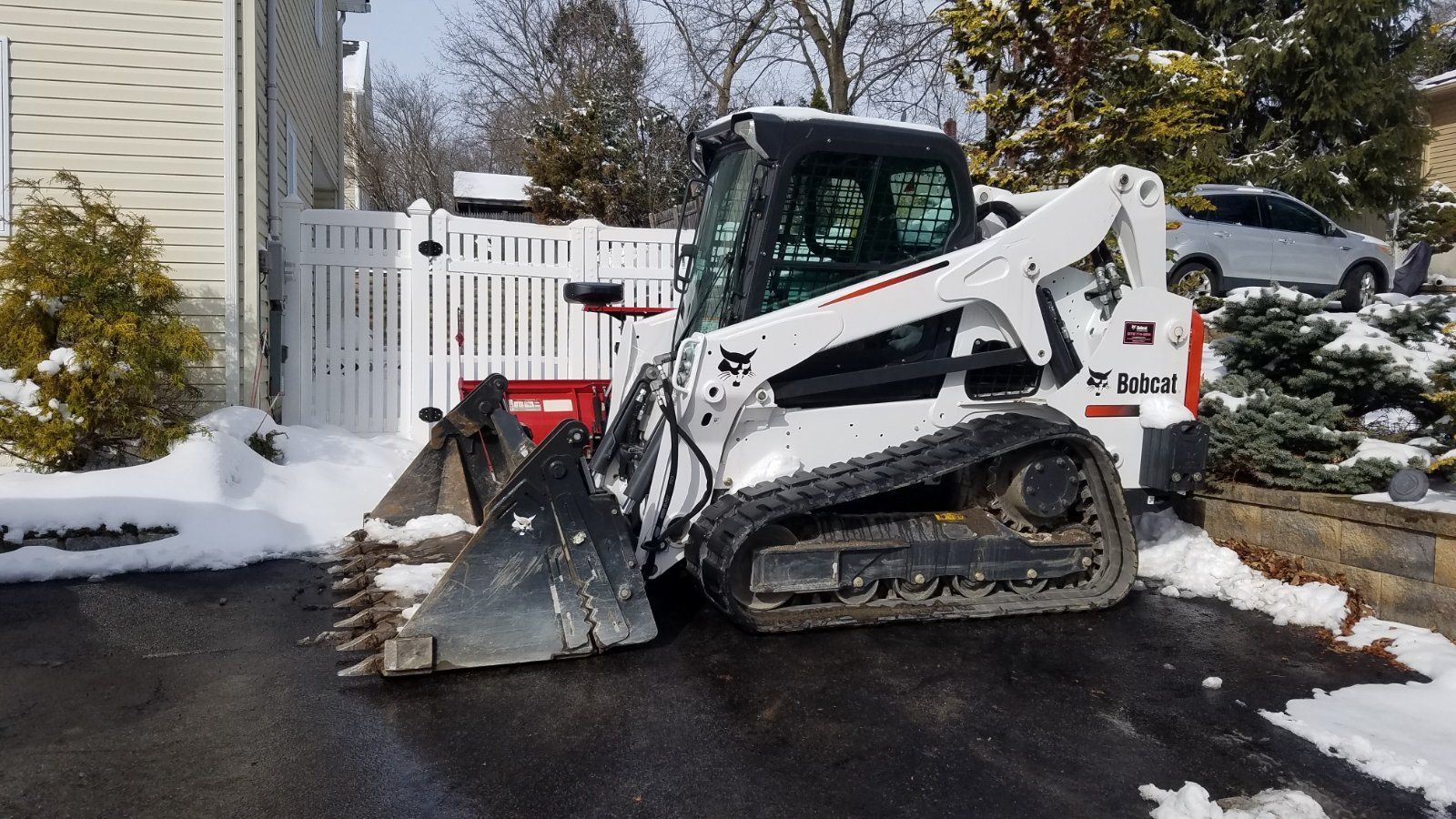 A white Bobcat track loader with a snow blade on a snow-covered driveway, near a white fence and house.