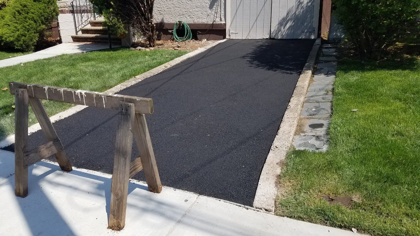 Newly paved black asphalt driveway bordered by concrete and grass, with a wooden sawhorse in the foreground.