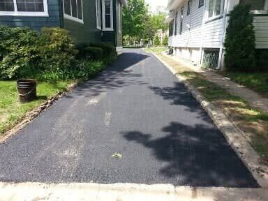 Newly paved asphalt driveway between two houses, trees casting shadows.