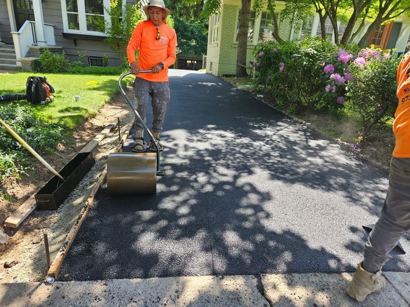 Workers paving a driveway with fresh asphalt. One man rolls a compactor, while another is partially visible.
