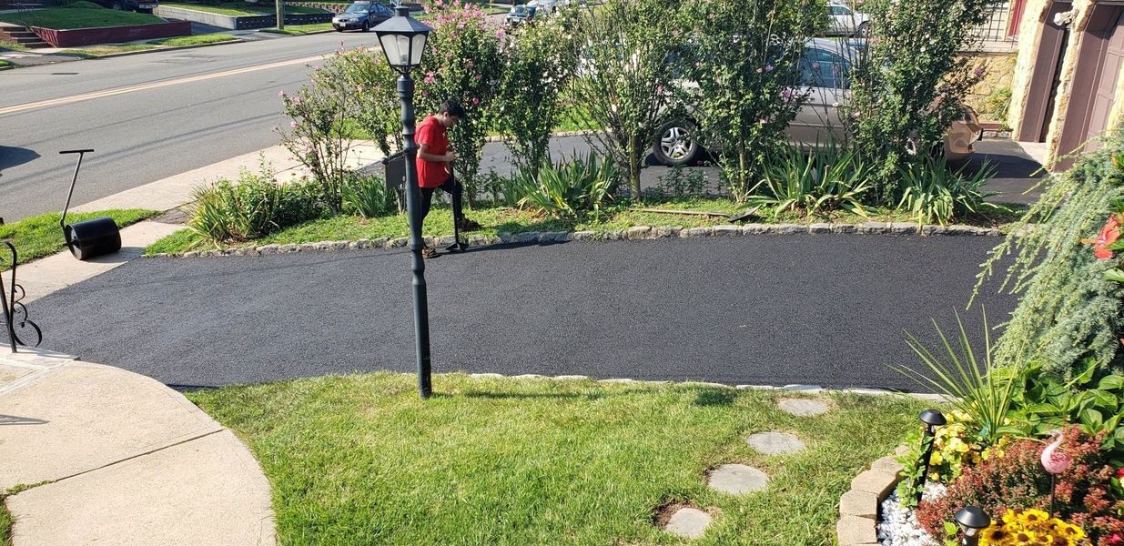A newly paved driveway, a grassy lawn, and a street are in the background. A person is seen near a mailbox.