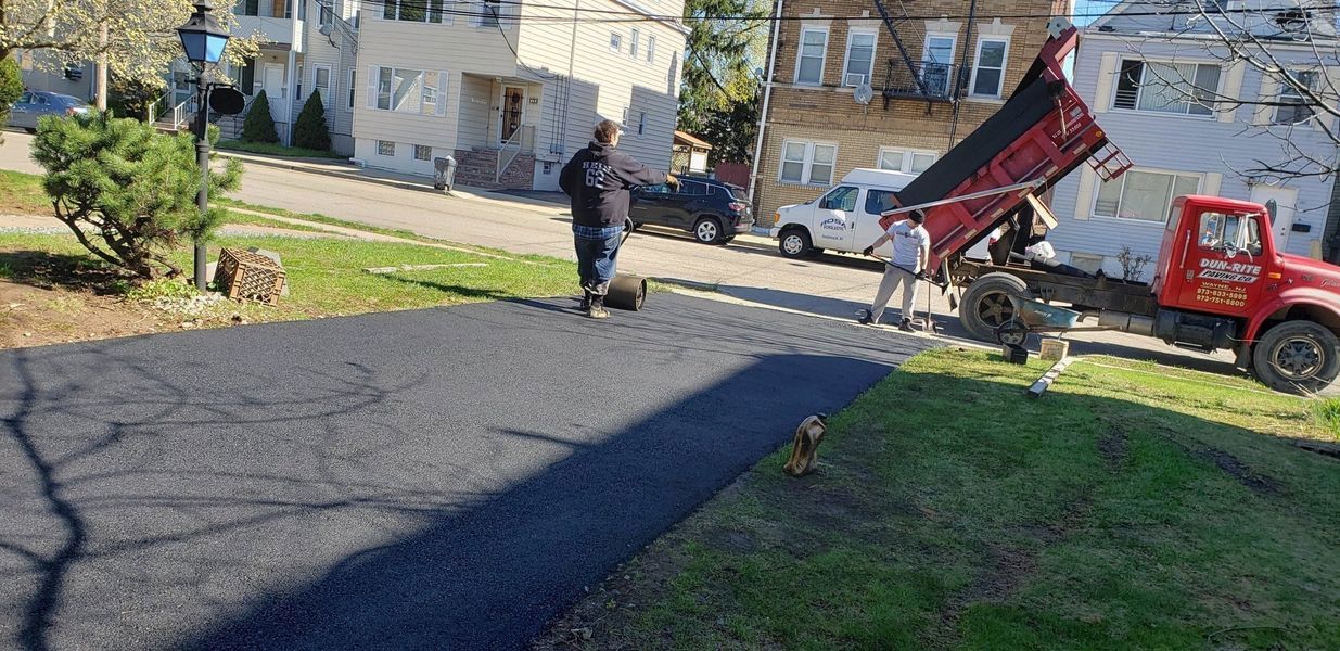 Workers paving a driveway with a dump truck. One man rolls the asphalt while another guides the process.