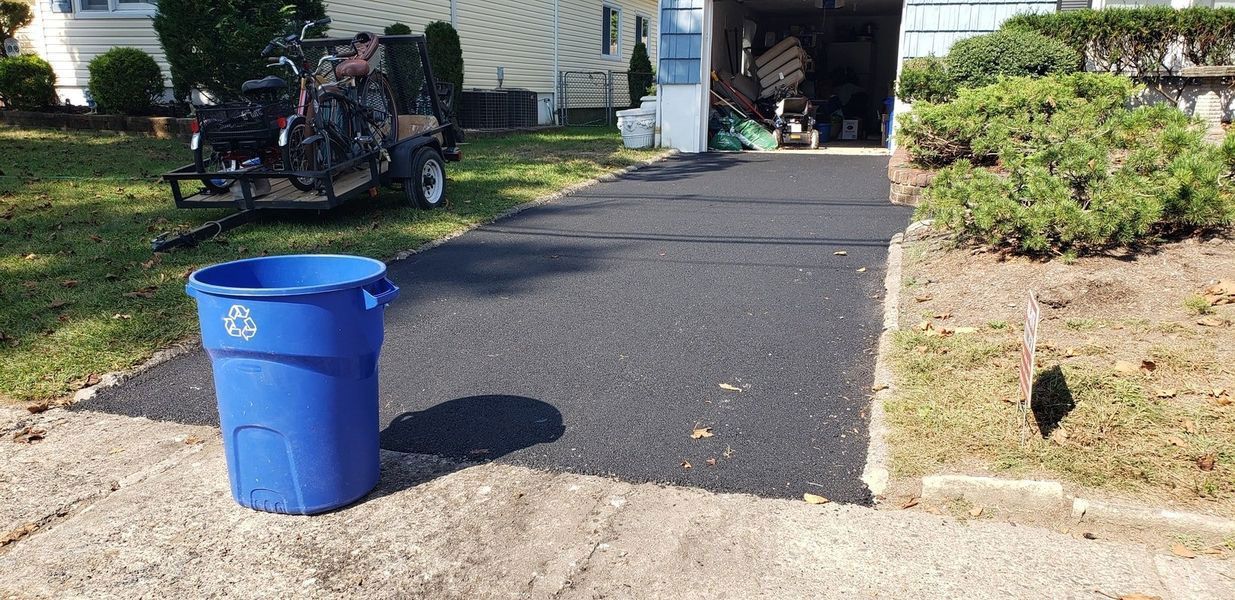 Blue recycling bin on a paved driveway, next to a garage and a grassy area with landscaping.
