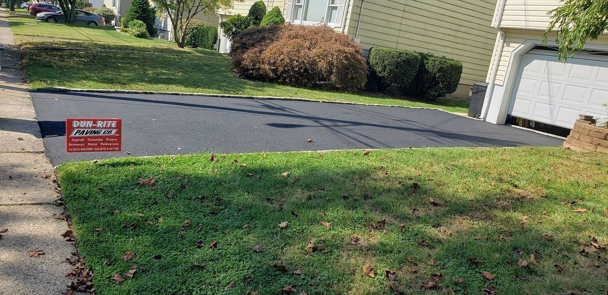 A driveway with fresh asphalt surrounded by green grass and a sign. In the background, a house and some bushes.