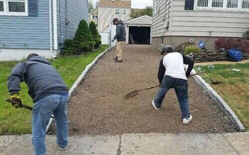 Three people spreading gravel on a driveway. One rakes, two shovel, all are in casual clothes on a sunny day.