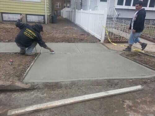Two workers pouring concrete for a sidewalk. One kneels, smoothing the surface; the other walks nearby.
