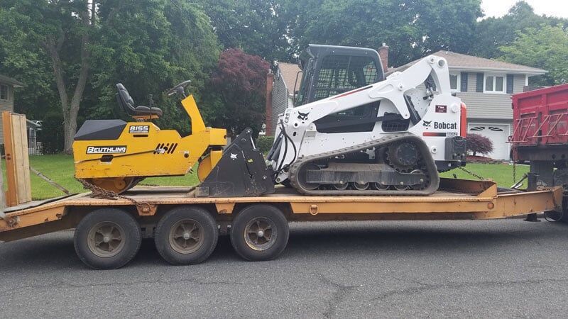 A yellow trailer carrying a yellow roller and a white Bobcat track skid steer loader parked on a residential street.