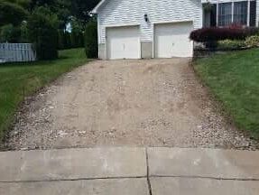 Gravel driveway leading to a white garage with two doors, in front of a house. Green grass surrounds.