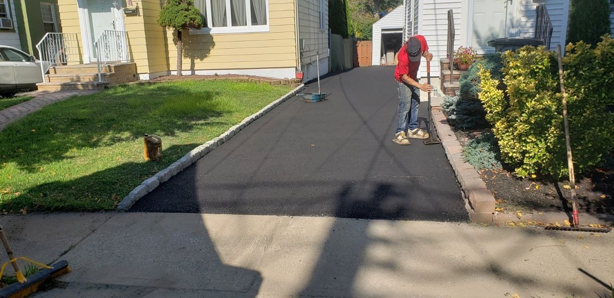 A person in a red shirt rakes a newly paved asphalt driveway. The driveway is flanked by grass and a garden.