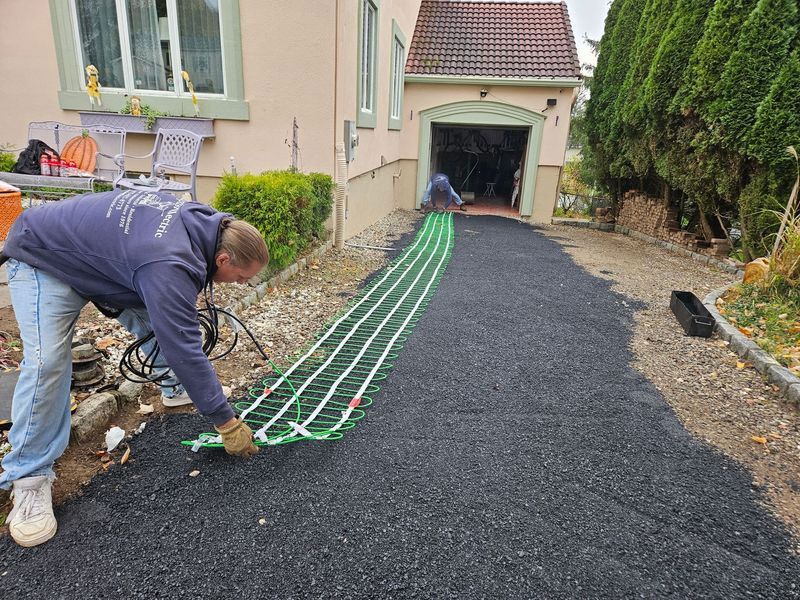 Two workers installing an electric heating system on a driveway covered with black asphalt in front of a house with a garage.