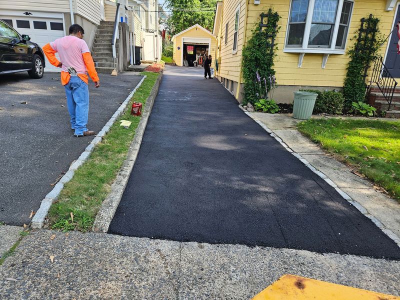 Freshly paved driveway next to a yellow house; a worker stands nearby, observing.