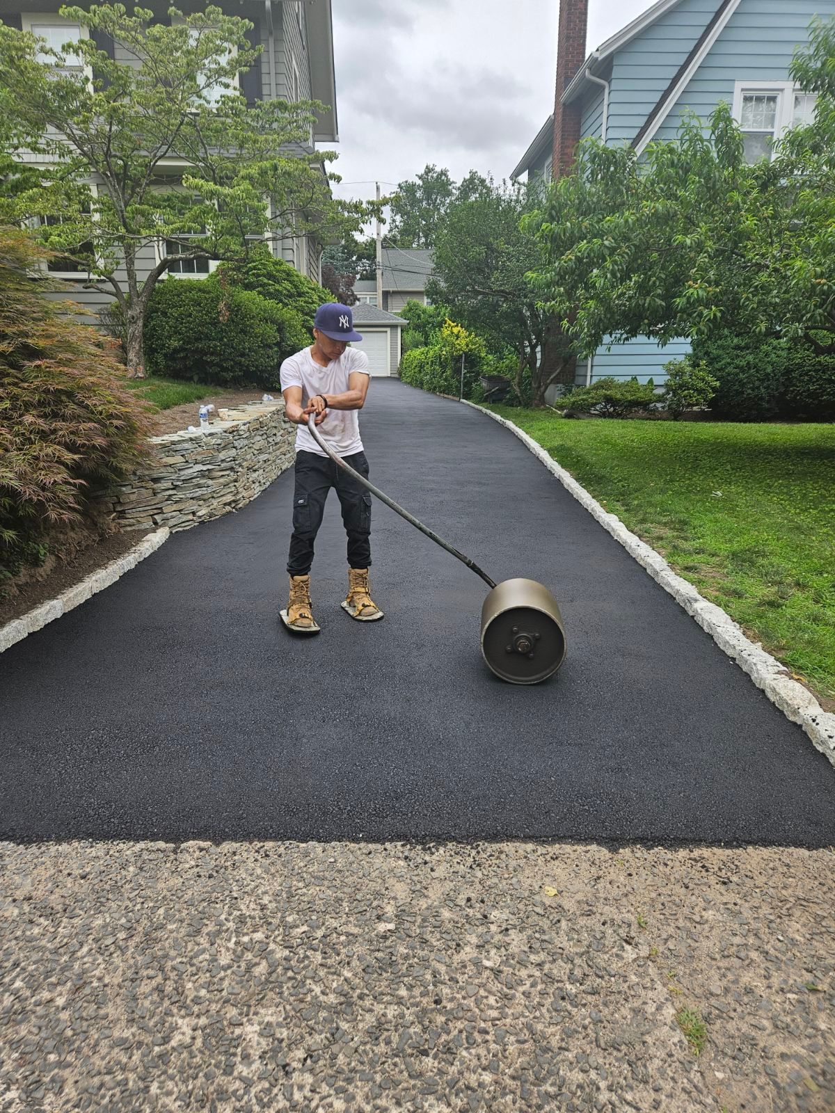 Person rolling a black compactor on a newly paved driveway in a residential neighborhood