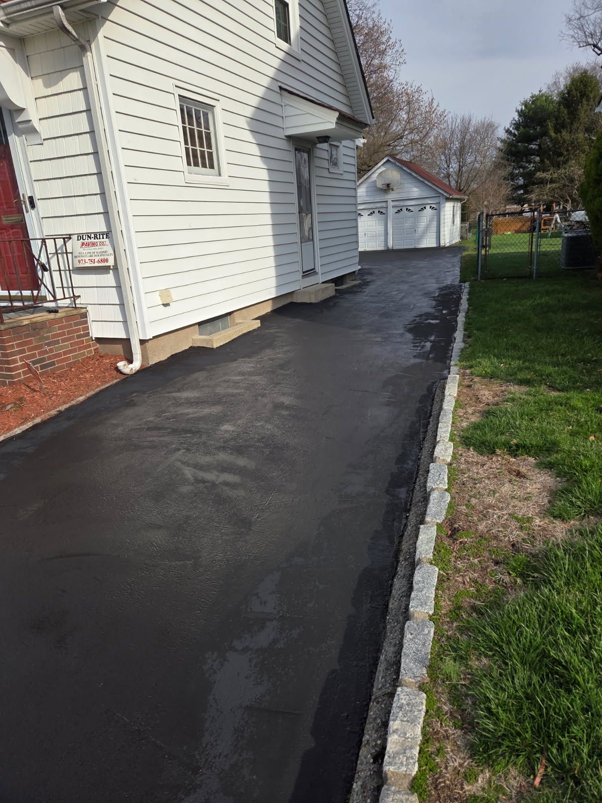 Freshly paved black asphalt walkway along the side of a white house with a brick foundation and grassy yard.