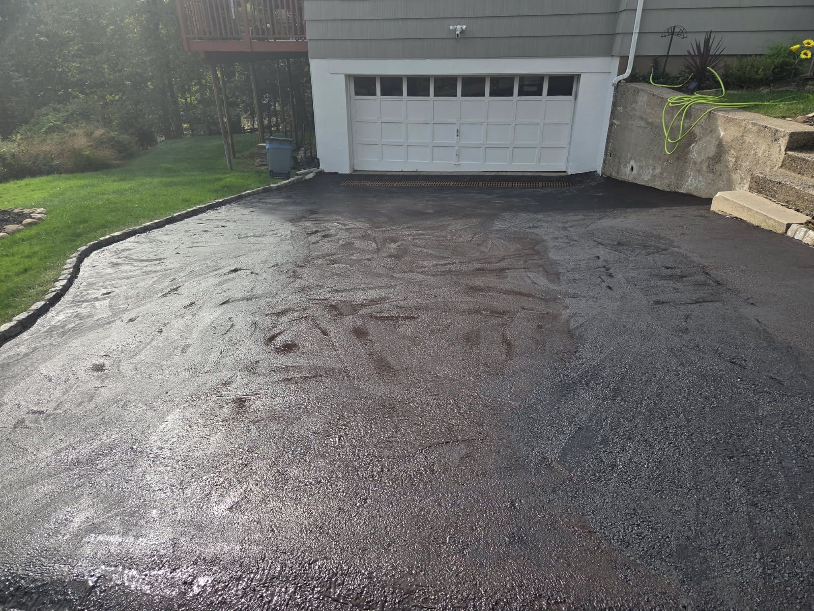 Wet driveway leading to a white garage door beside a house and stone retaining wall.