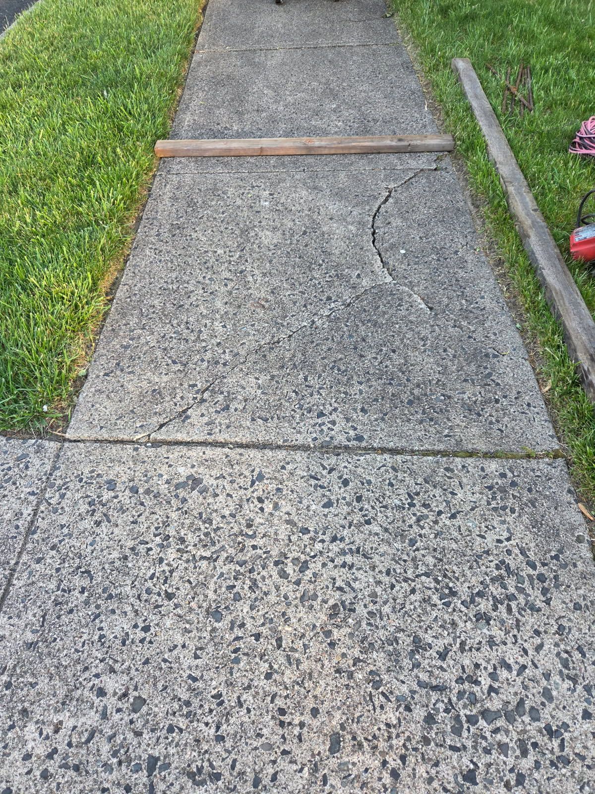 Cracked concrete sidewalk with grass borders and a wooden plank across one section