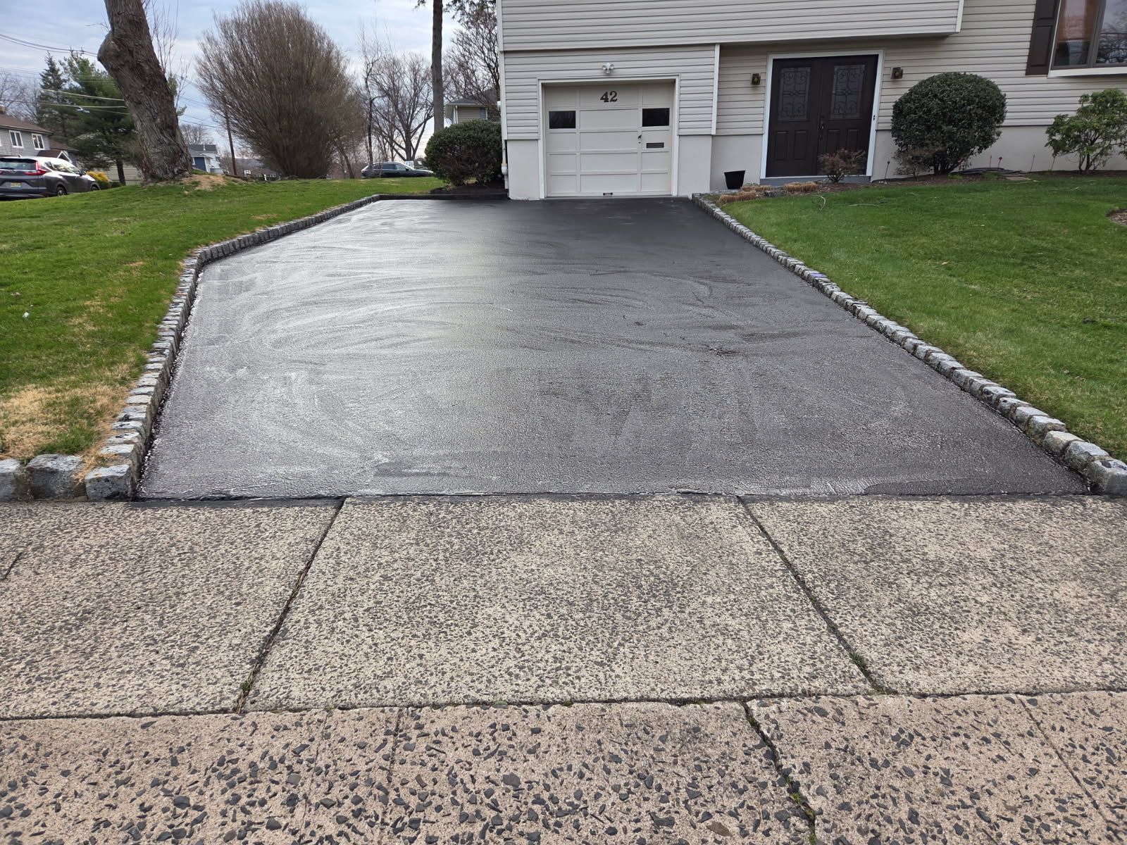Freshly paved black driveway leading to a house garage, bordered by grass and a sidewalk