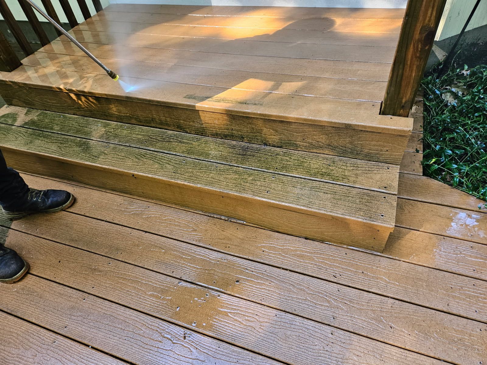 Wet wooden deck and glass railing beside black boots and a doorway reflected in sunlight