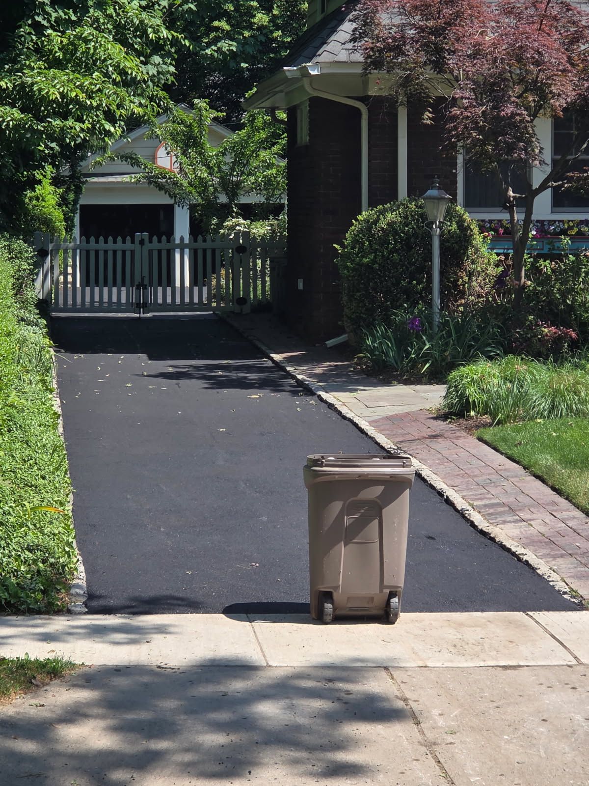 Trash bin on a driveway in front of a house and gate, with trees and shrubs lining the path.