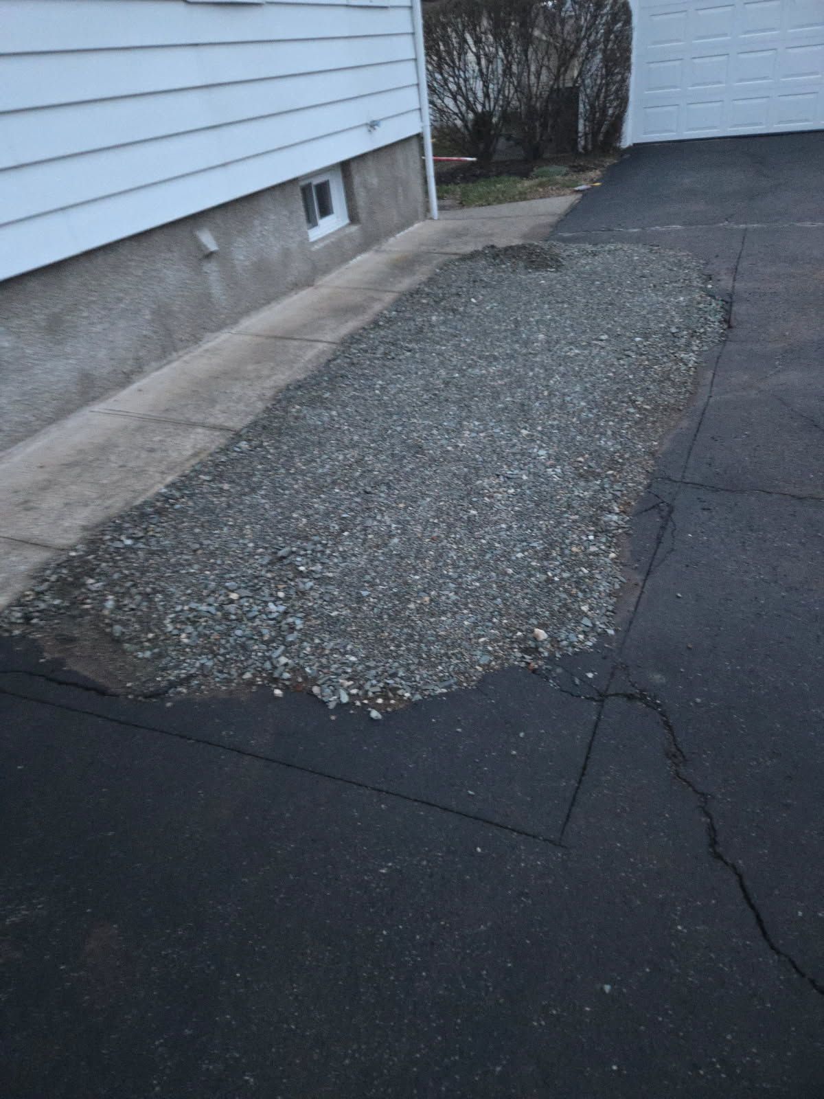 Cracked asphalt driveway beside a white house with a gravel patch near the foundation.