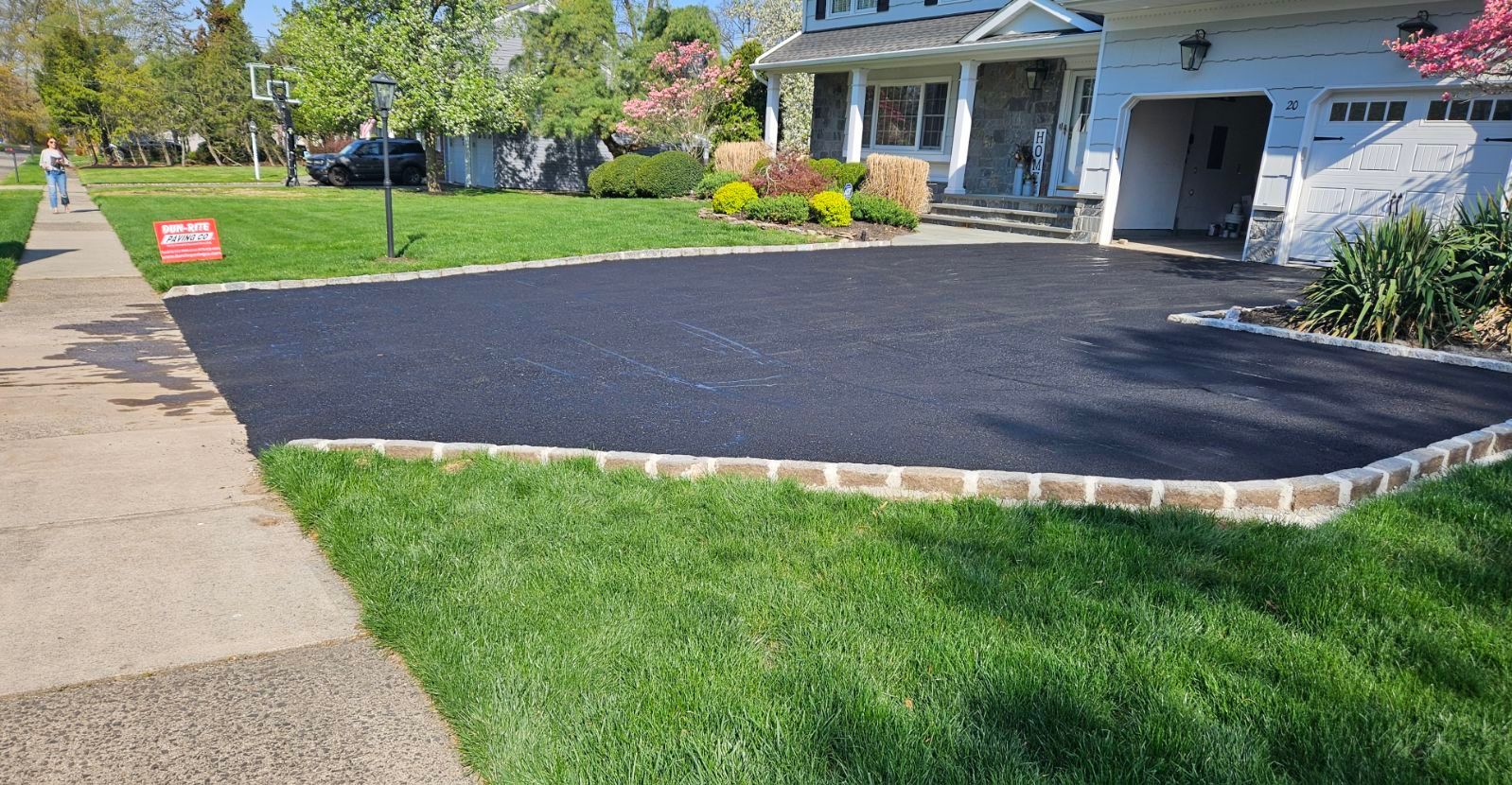 Residential driveway with fresh black asphalt, stone edging, and a house front yard on a sunny day