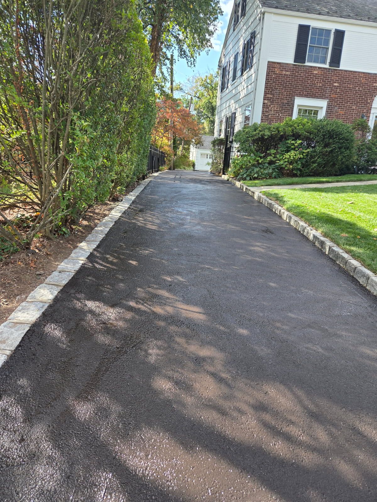 Paved driveway beside a brick house, lined with trees and green lawn in bright sunlight.