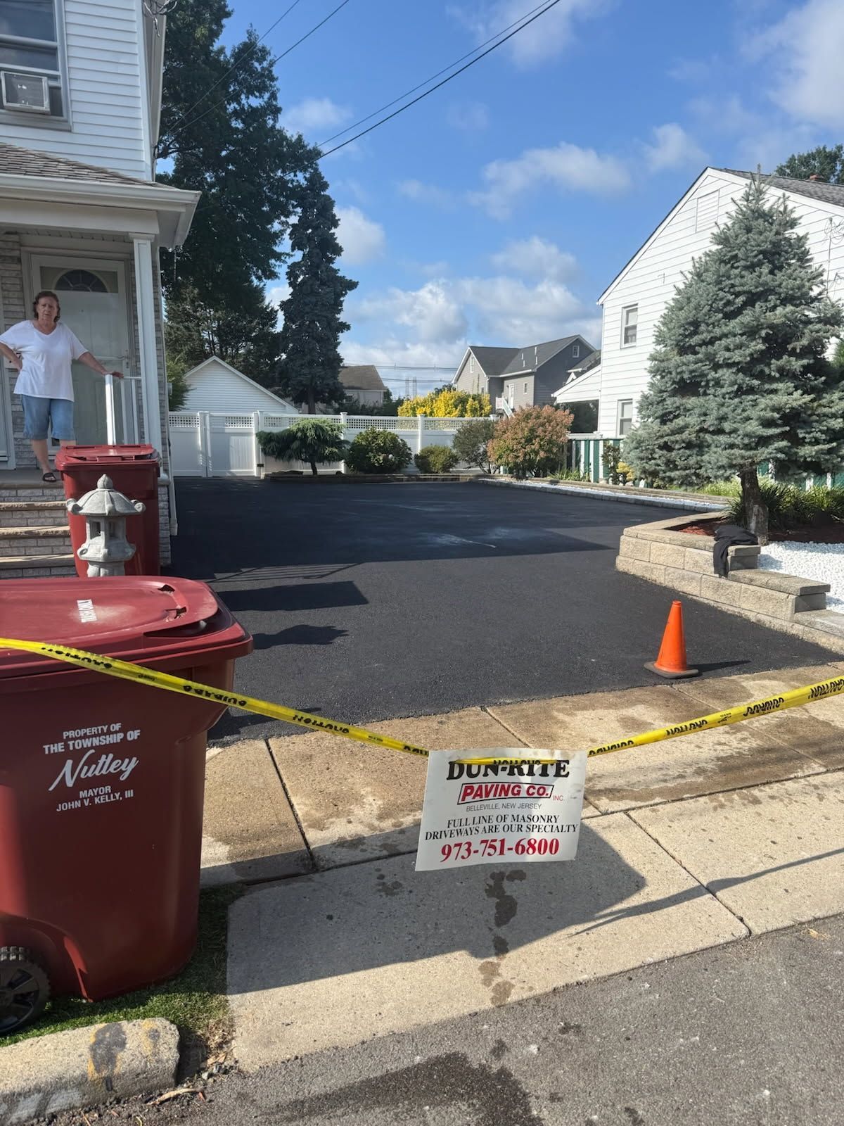 Driveway with yellow caution tape, red trash bin, orange cone, and a sign for a dumpster service.