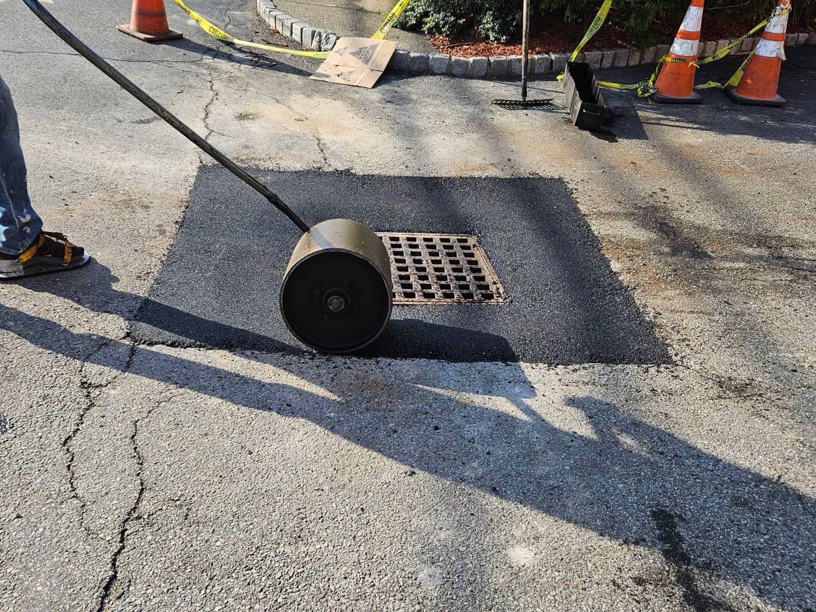 Roadwork scene with a worker rolling a black pavement roller beside a storm drain and orange cones.