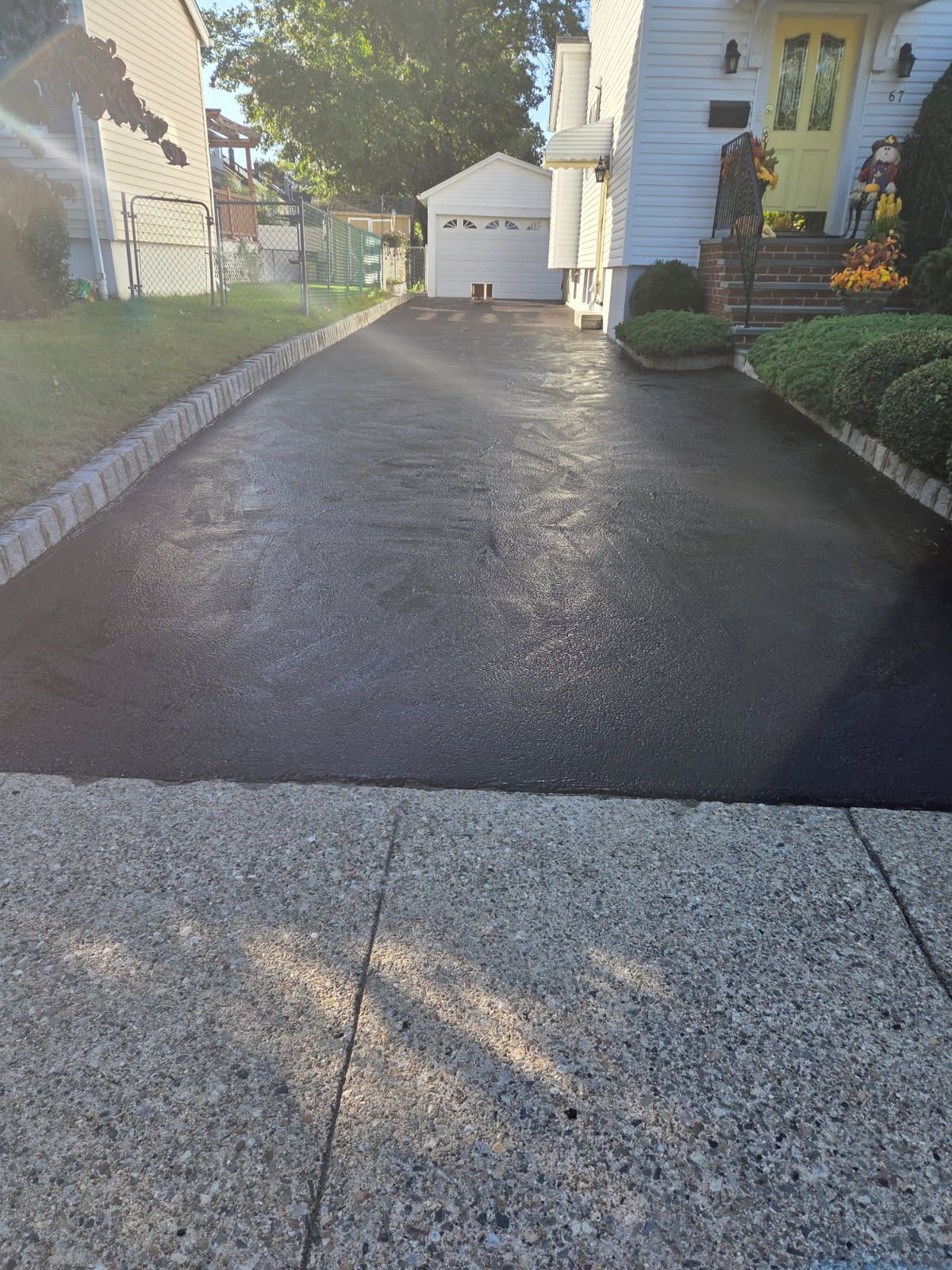 Wet asphalt driveway between houses, with sunlight and flower beds along the sides