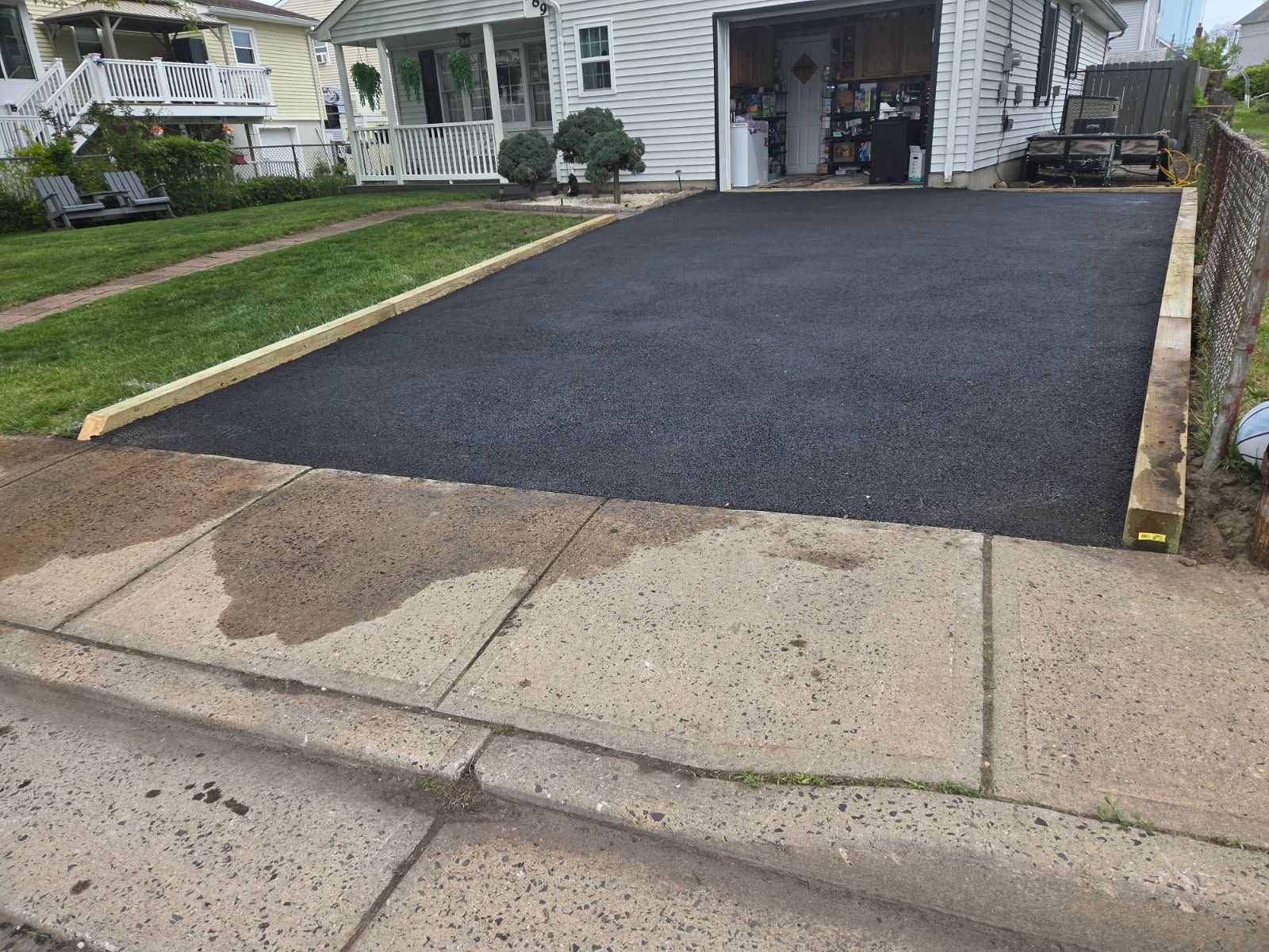 Fresh black asphalt driveway beside a house, bordered by curb and grass, with a garage in the background