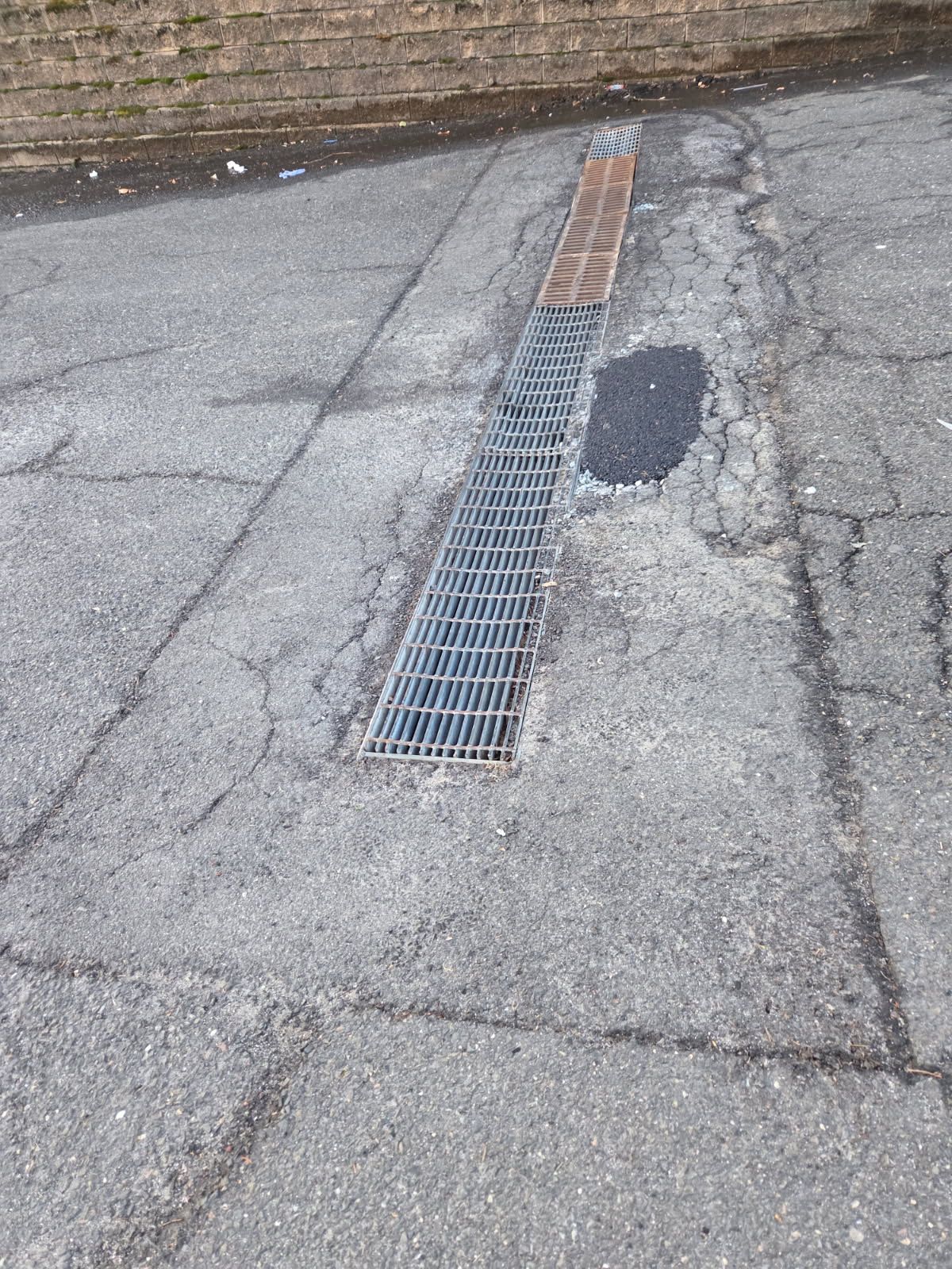 Cracked asphalt road with a long metal drain grate and a small repair patch near the curb