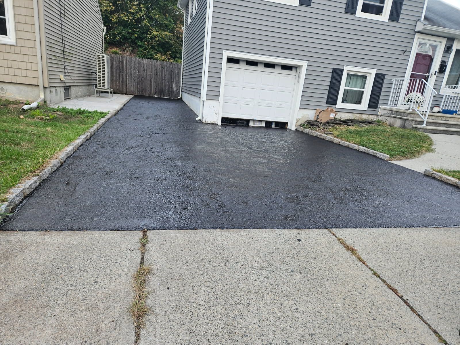 Fresh black asphalt driveway leading to a gray house with a white garage door and side yard.