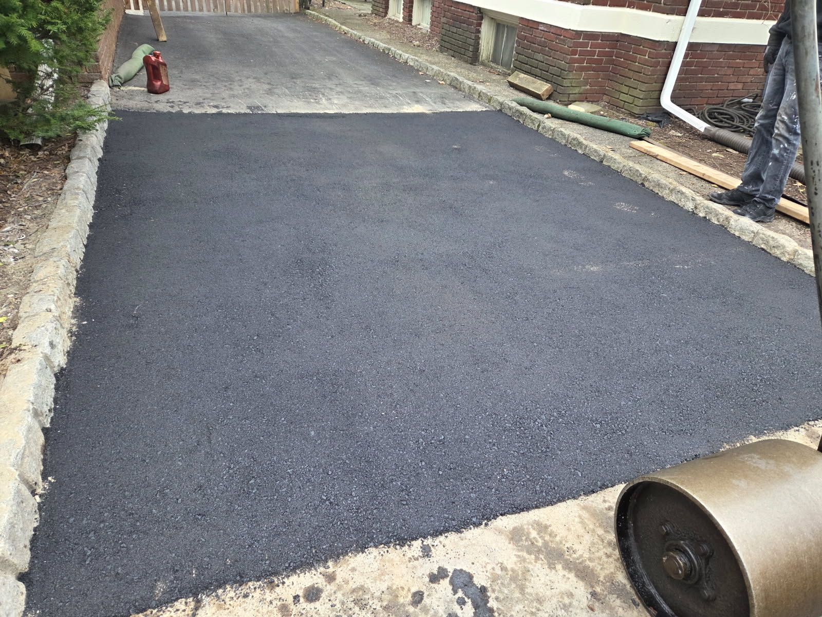 Fresh black asphalt paving on a driveway beside brick walls and a curb, with a roller in the foreground