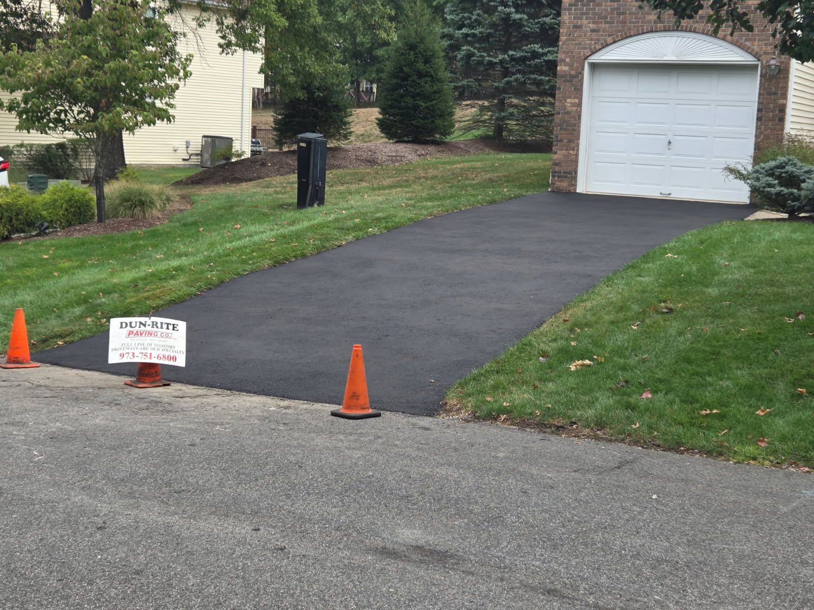 Freshly paved black driveway with orange cones and a small sign in front of a white garage