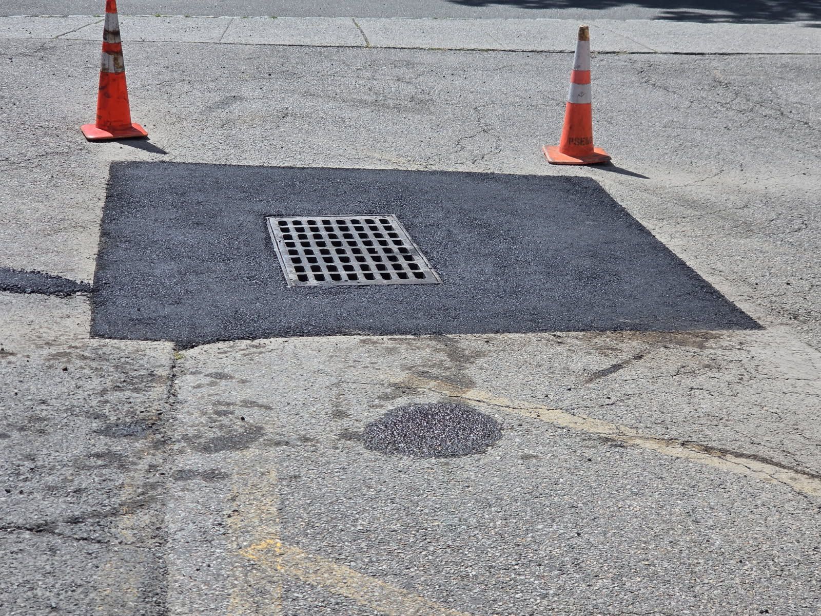 Road repair patch with a metal drain grate, flanked by two orange traffic cones.