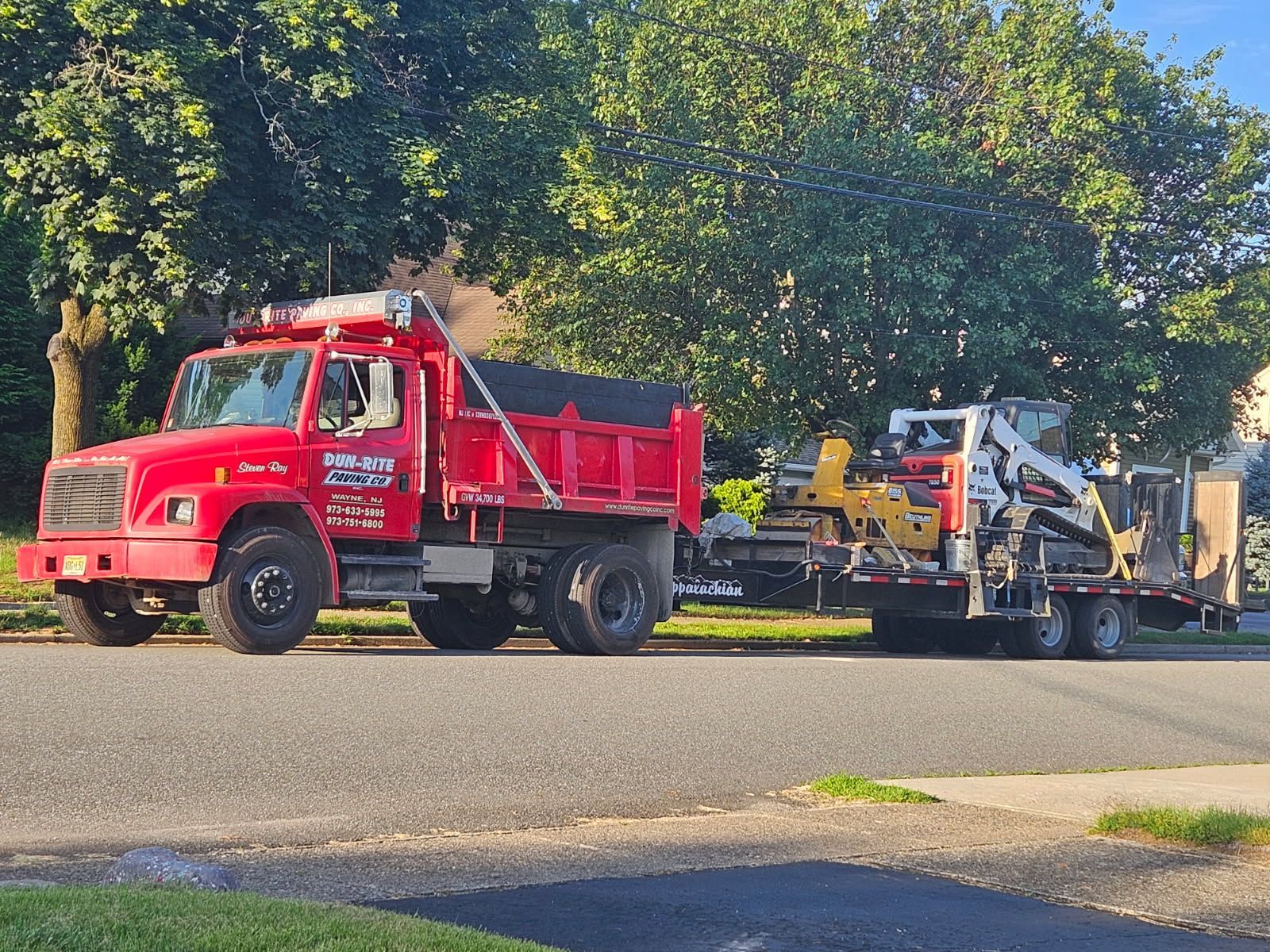 Red tow truck hauling a yellow utility vehicle on a trailer along a tree-lined street.