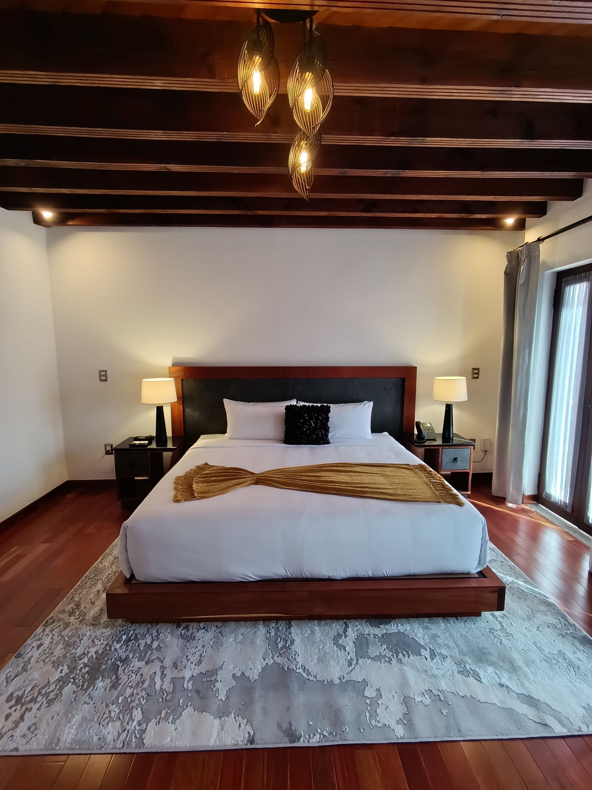 Dormitorio con cama, mesitas de noche, alfombra y lámparas de techo decorativas. Suelo y vigas de madera.