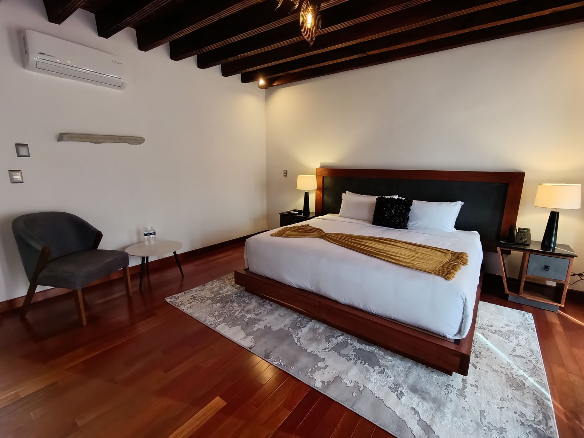 Dormitorio con suelo de madera, cama, silla y mesitas de noche; paredes blancas y techo con vigas.