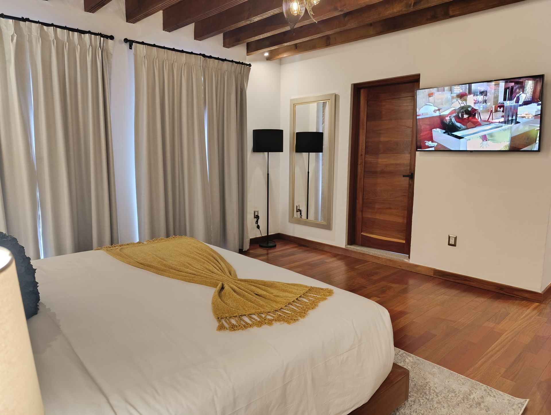 Dormitorio con cama, TV, suelo de madera, cortinas color crema y espejo.