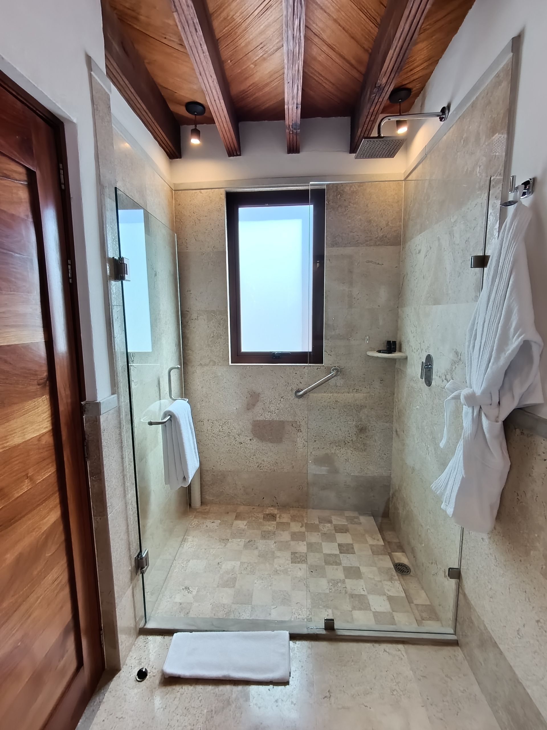 Baño con paredes de piedra, techo de madera, mamparas de cristal y ventana. Hay una bata colgada en la pared derecha.