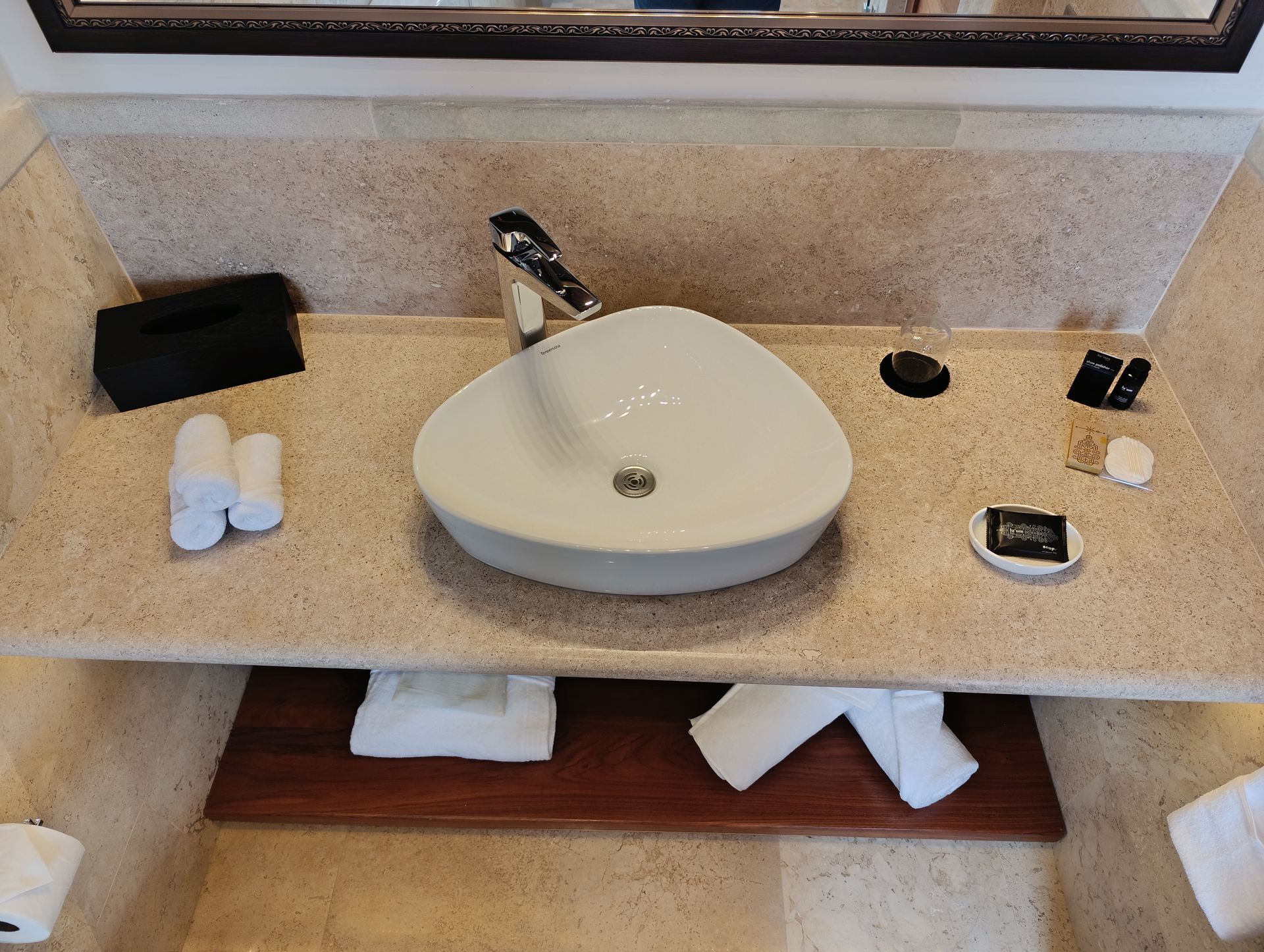 Tocador de baño con lavabo blanco, grifo negro y varios artículos de tocador sobre una encimera beige.