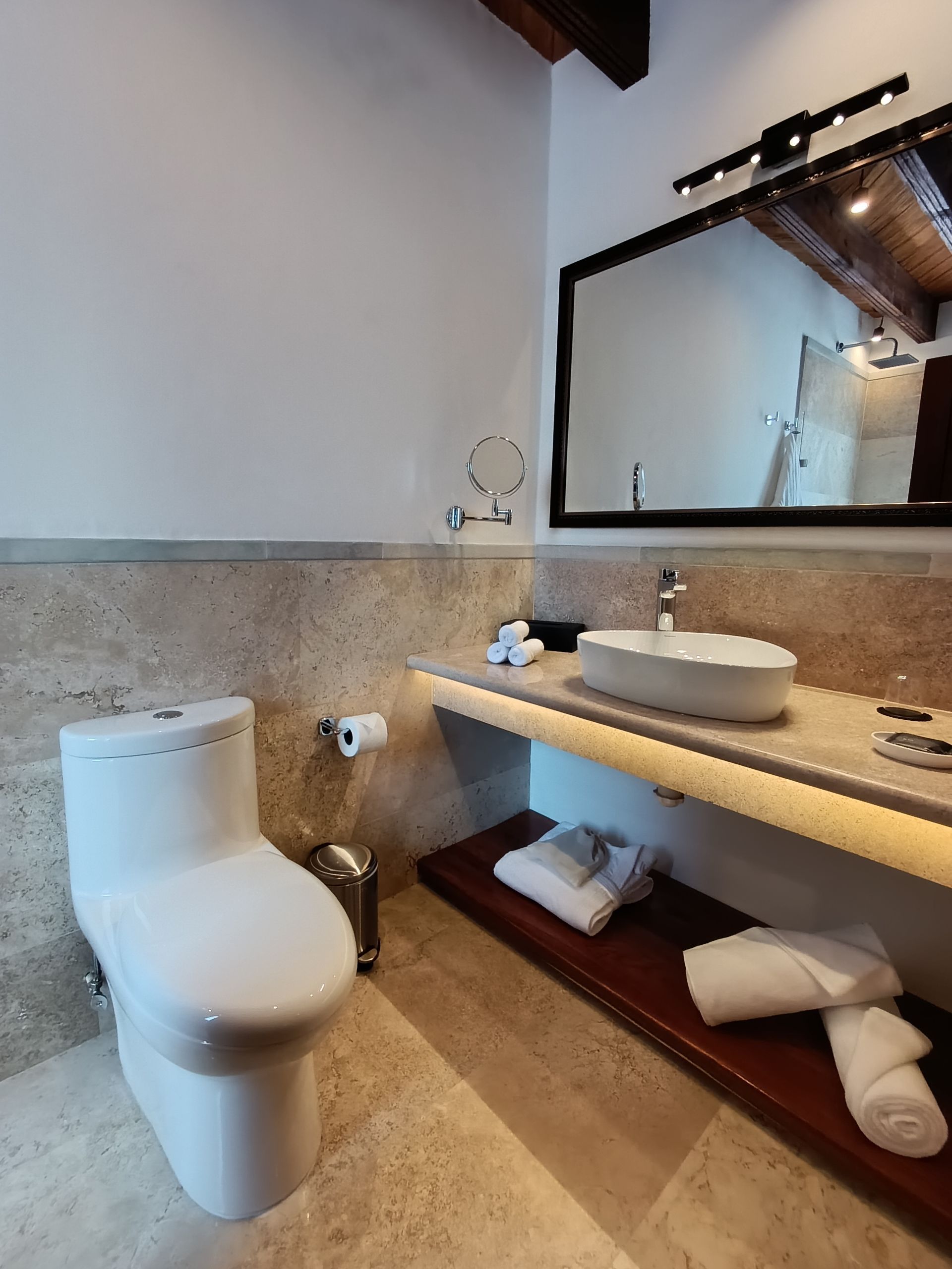 Baño con inodoro blanco, lavabo sobre encimera de mármol, gran espejo y techo de madera.