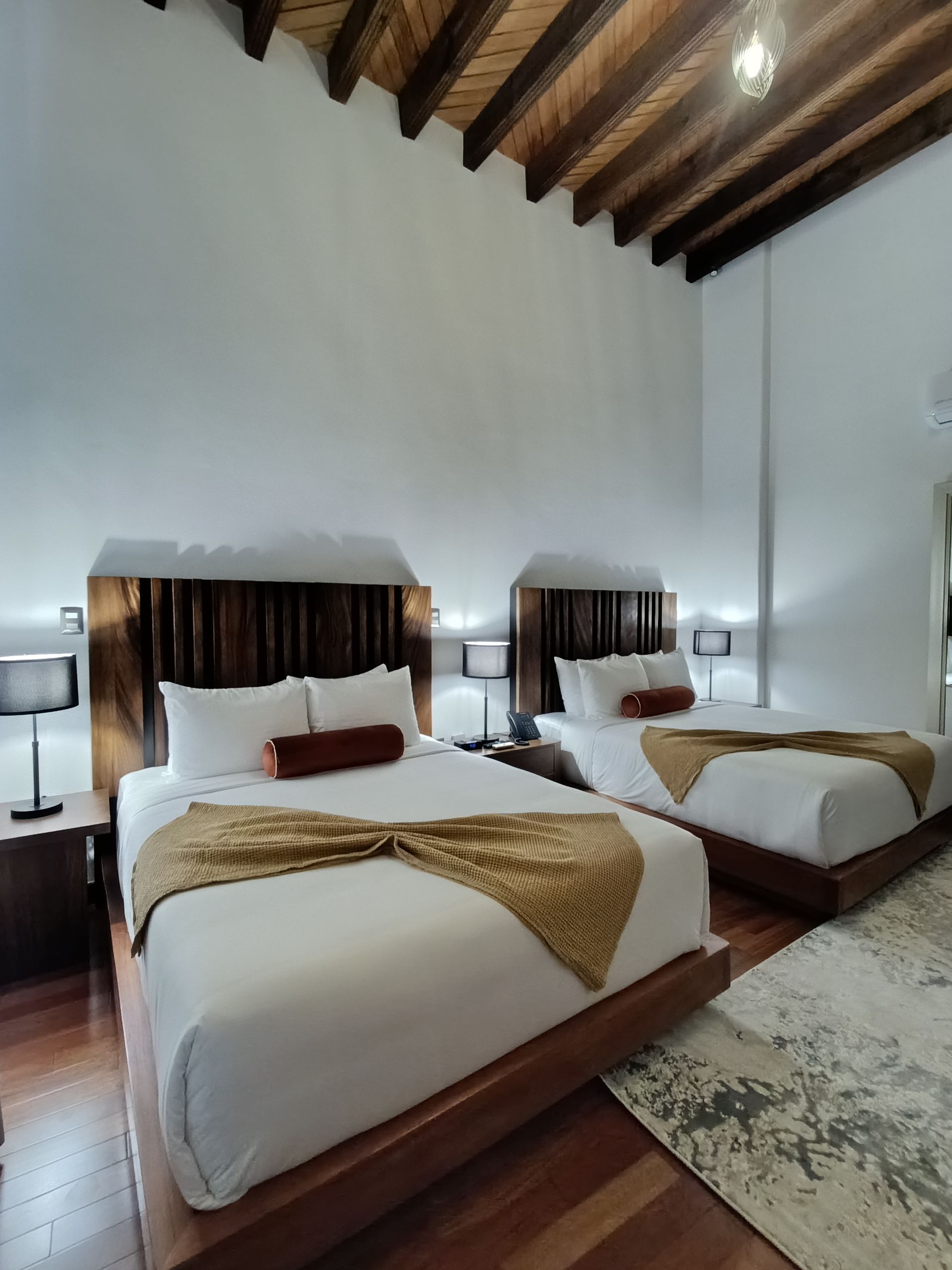 Dos camas en una habitación con techo de madera y pisos de madera, cada una con sábanas blancas y mantas doradas.