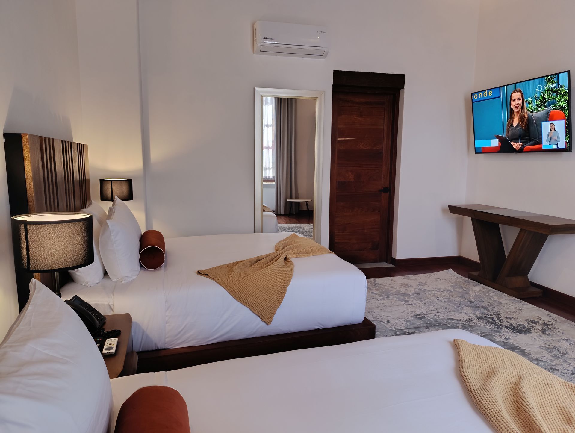 Habitación de hotel con dos camas, televisor y puerta de madera. Paredes blancas, molduras de madera y alfombra gris.