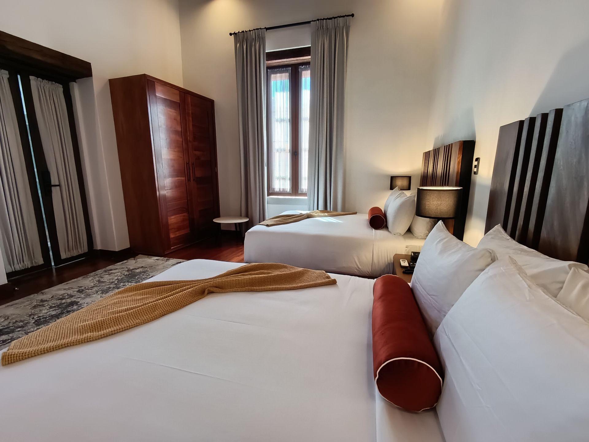Dormitorio con dos camas, armario de madera y ventana con cortinas. Ropa de cama blanca y almohadas naranjas y rojas.