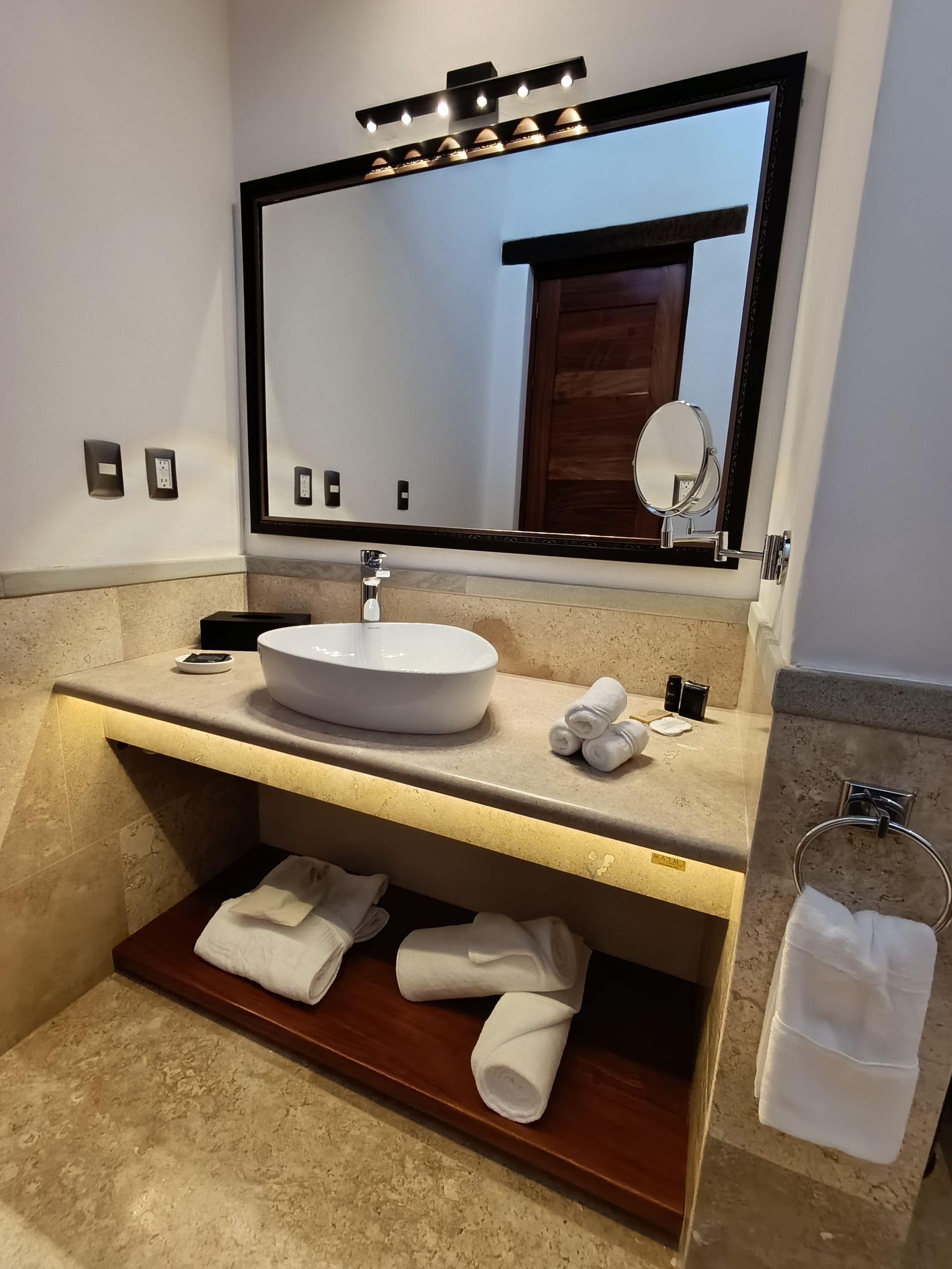 Baño con lavabo de color blanco, espejo grande y estante abierto con toallas.