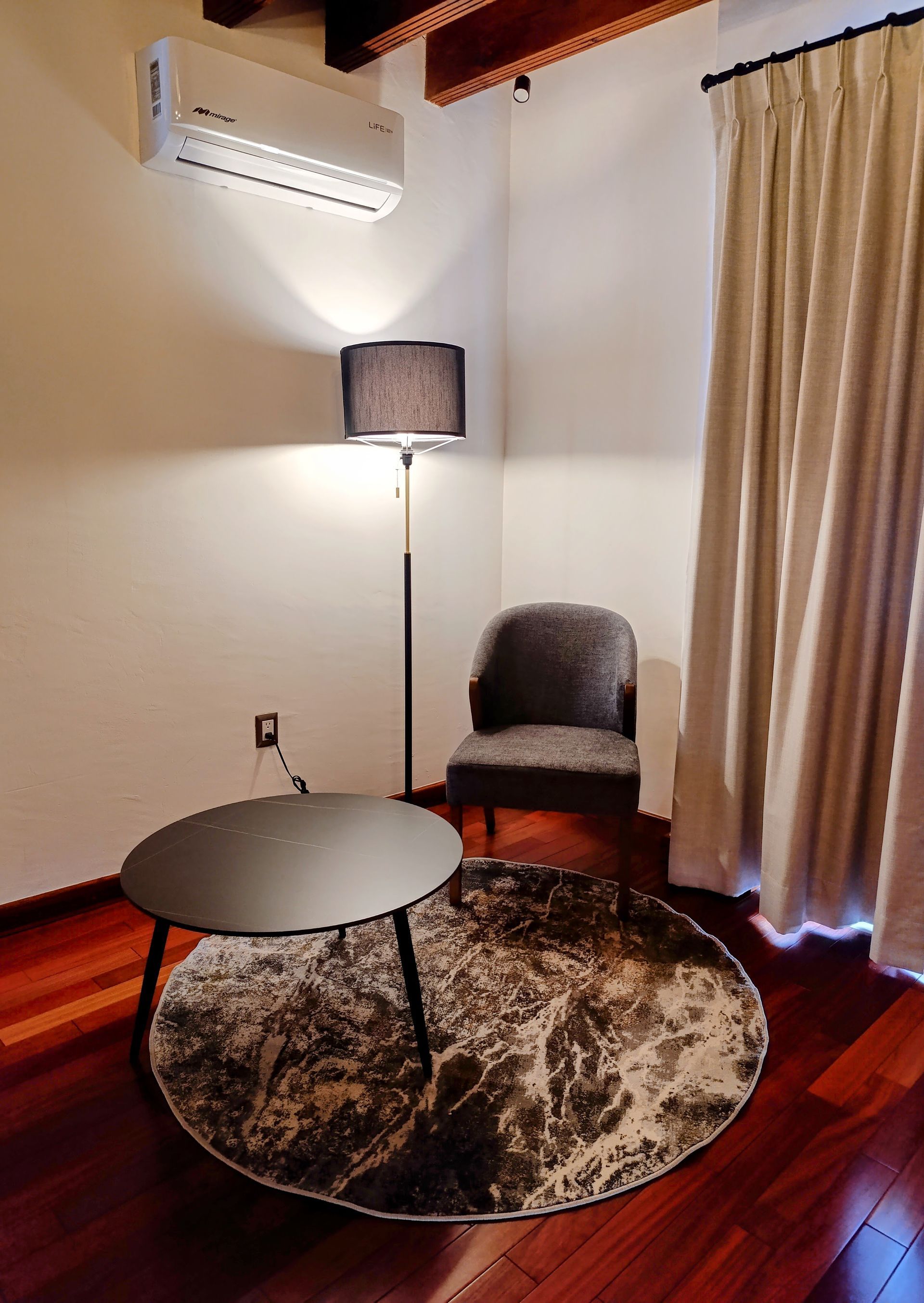 Rincón acogedor: silla, lámpara, mesa de centro redonda y alfombra sobre suelo de madera, con aire acondicionado y cortinas.