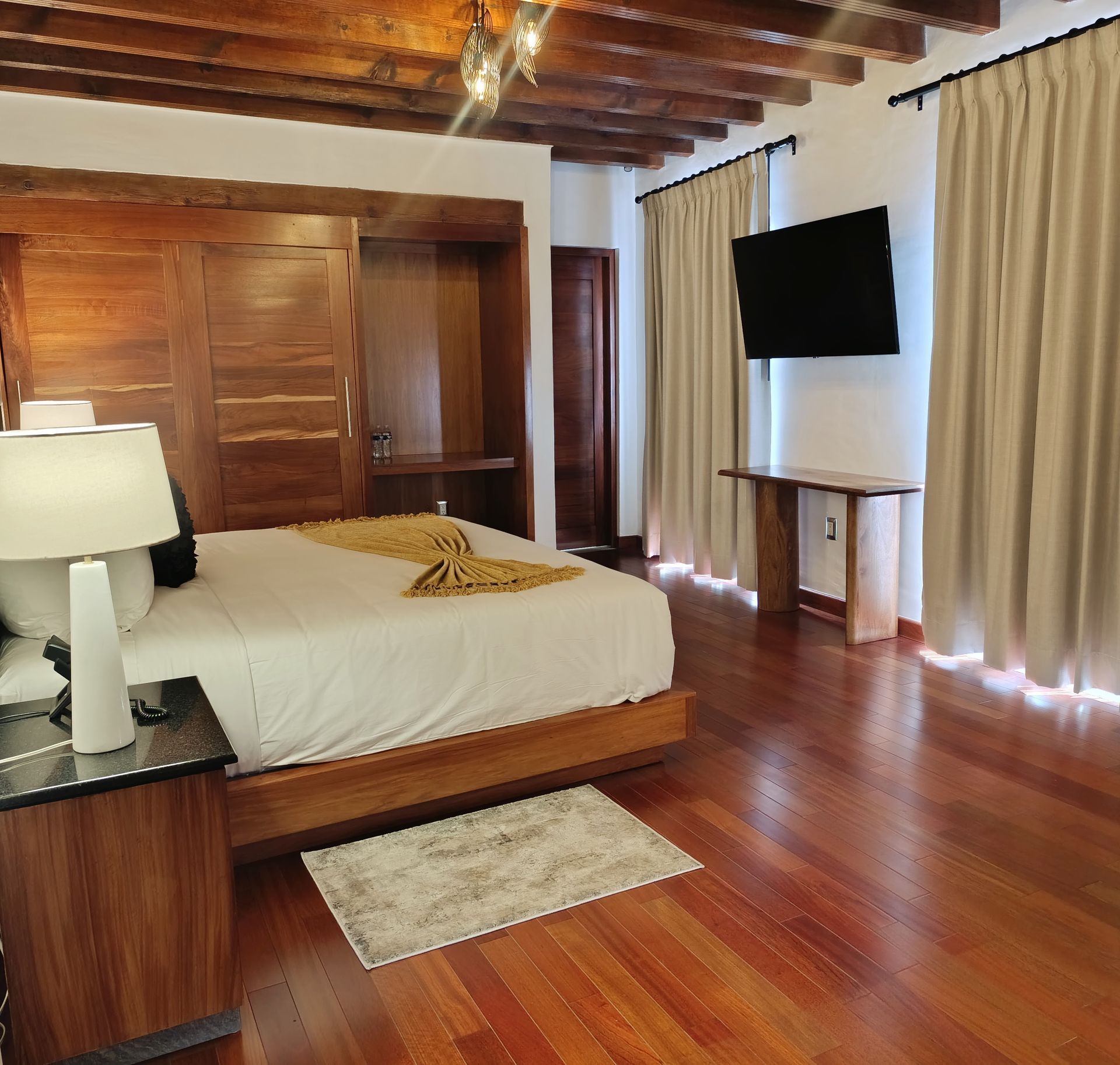 Dormitorio con muebles de madera, cama, TV y cortinas color crema.