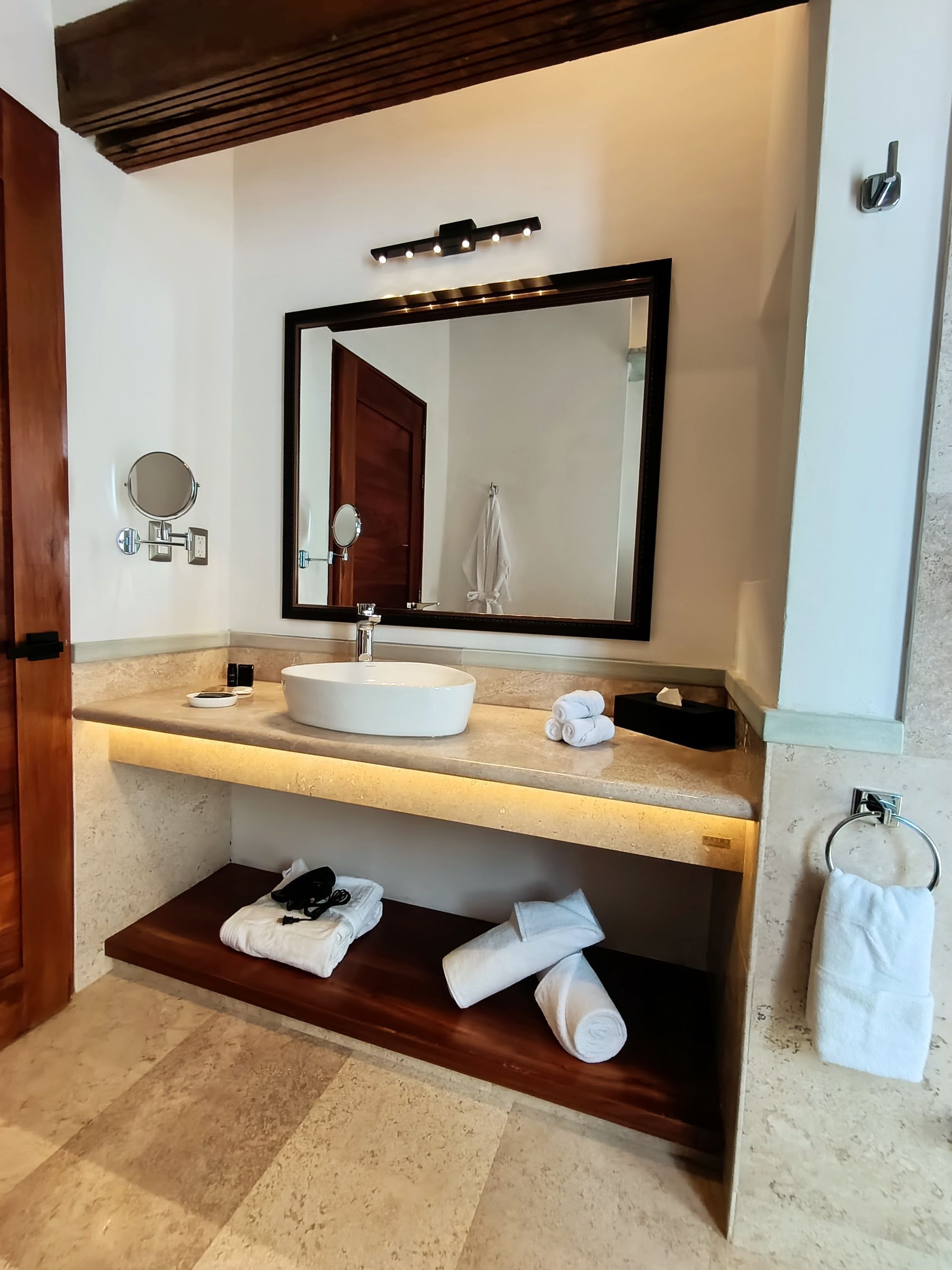 Baño con lavabo blanco, espejo de marco oscuro, detalles de madera y toallas apiladas.
