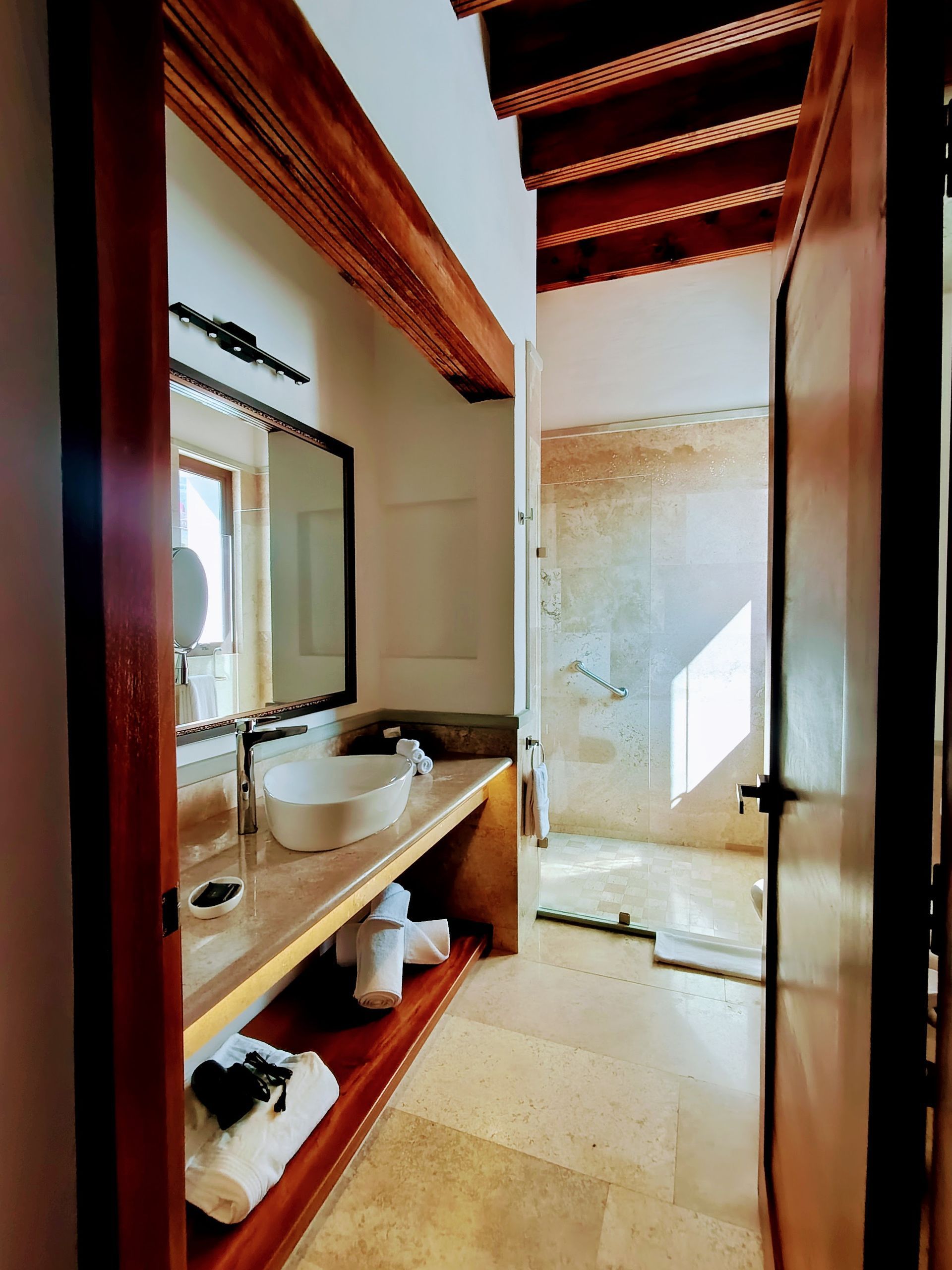 Baño con vigas de madera, tocador y zona de ducha visible a través de una puerta abierta.
