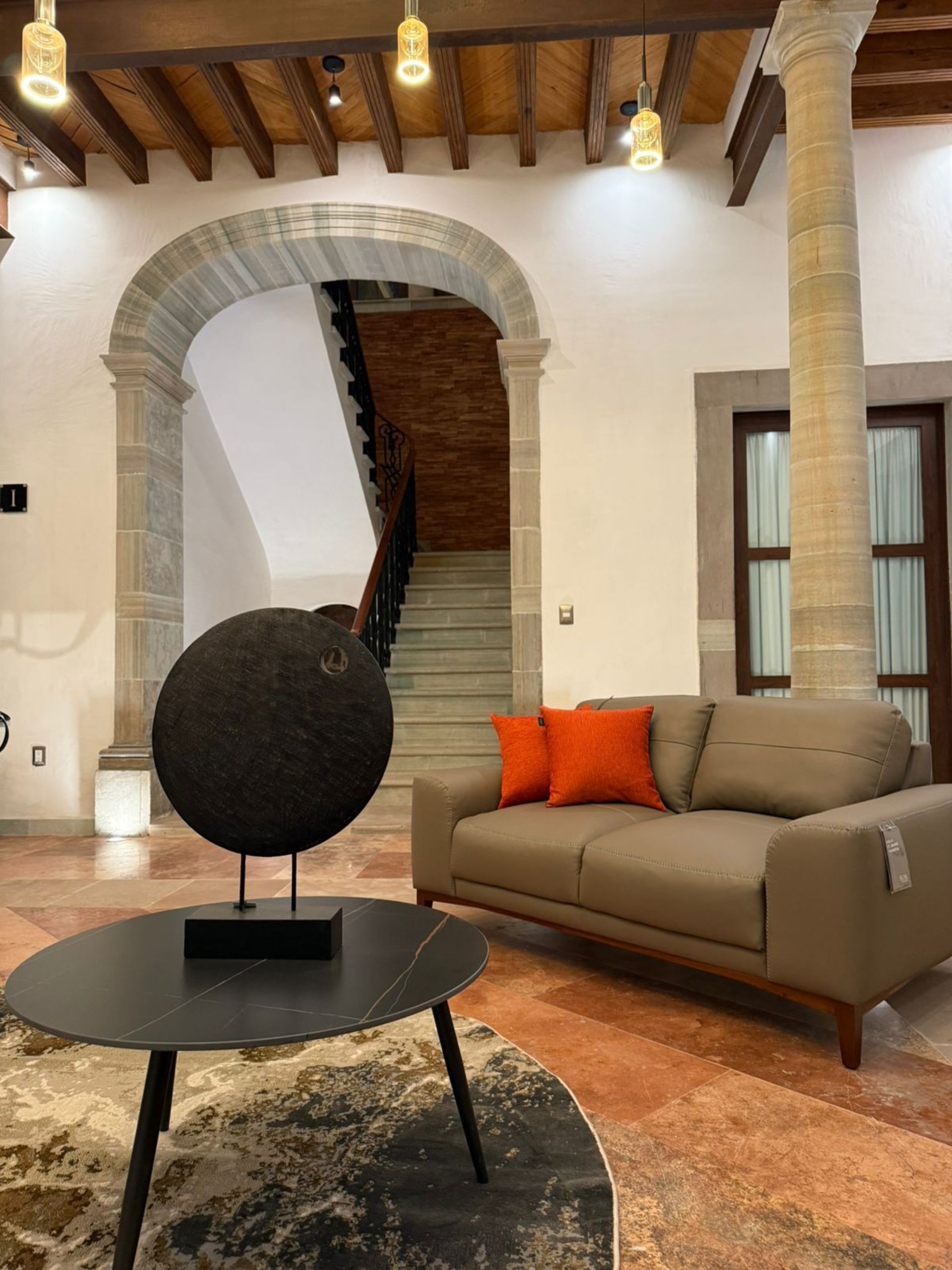 Sala de estar con un sofá marrón, almohadas naranjas y una pieza de arte negra, escalera al fondo.